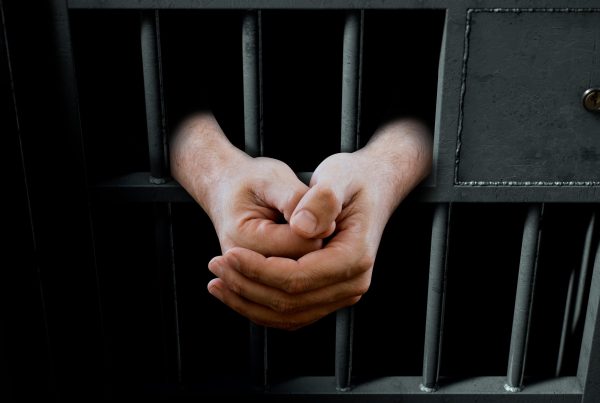 A closeup of a dimly lit prison holding cell door with arms reaching out in a folded position